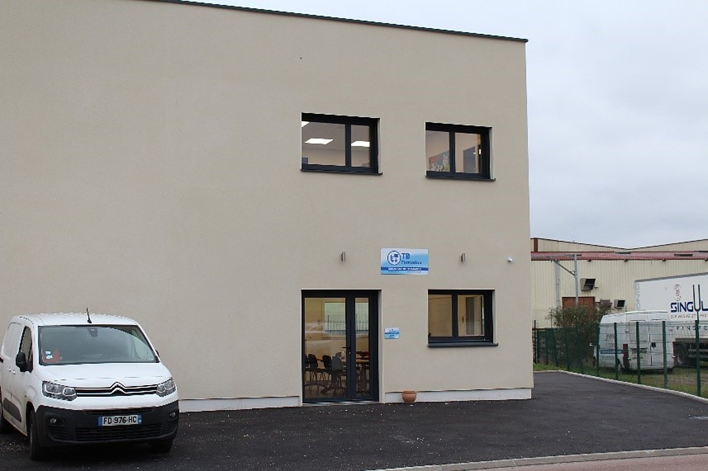 Bâtiment moderne de couleur beige avec une enseigne bleue près de la porte vitrée et une fourgonnette blanche garée sur un parking en enrobé.
