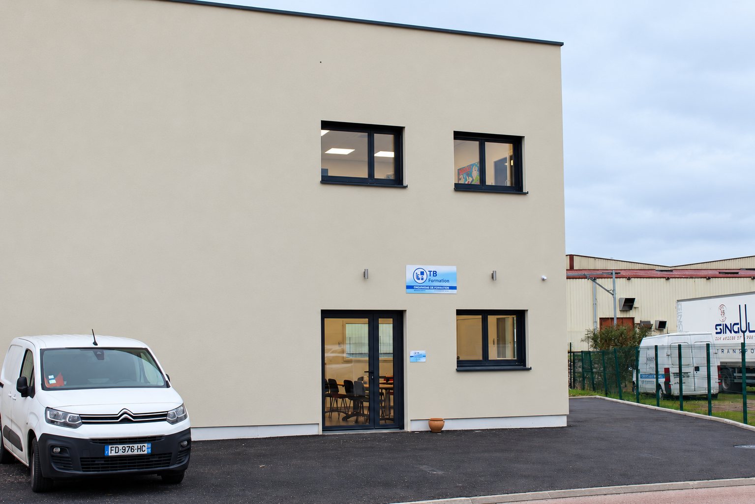 Bâtiment beige à toit plat portant l’enseigne TB Formation, avec une camionnette blanche garée sur le parking en enrobé devant l’entrée vitrée.
