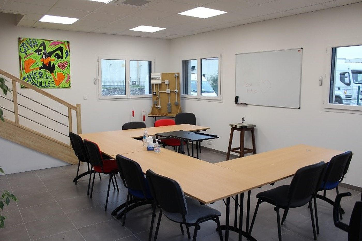 Une salle de formation moderne avec des tables disposées en U, des chaises noires, un tableau blanc, un panneau électrique pédagogique et un tableau coloré au mur.