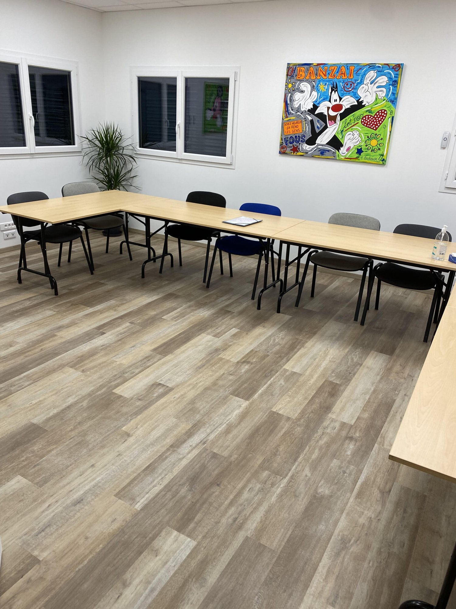 Salle de réunion lumineuse avec des tables en bois disposées en U, des chaises variées, un parquet clair et un tableau pop art coloré au mur.